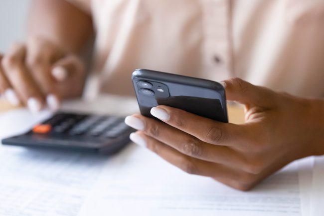 A person sits at a table with papers spread out, holding a smartphone in one hand and using a calculator with the other.
