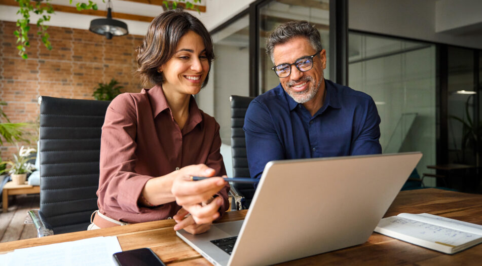 Two people seated at a wooden table in an office, reviewing content on a laptop.