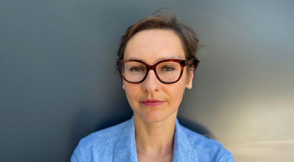 A person wearing glasses and a blue shirt stands against a plain dark background. The face is blurred and no specific details or text are visible.