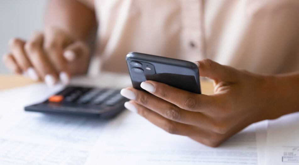 A person sits at a table with papers spread out, holding a smartphone in one hand and using a calculator with the other.