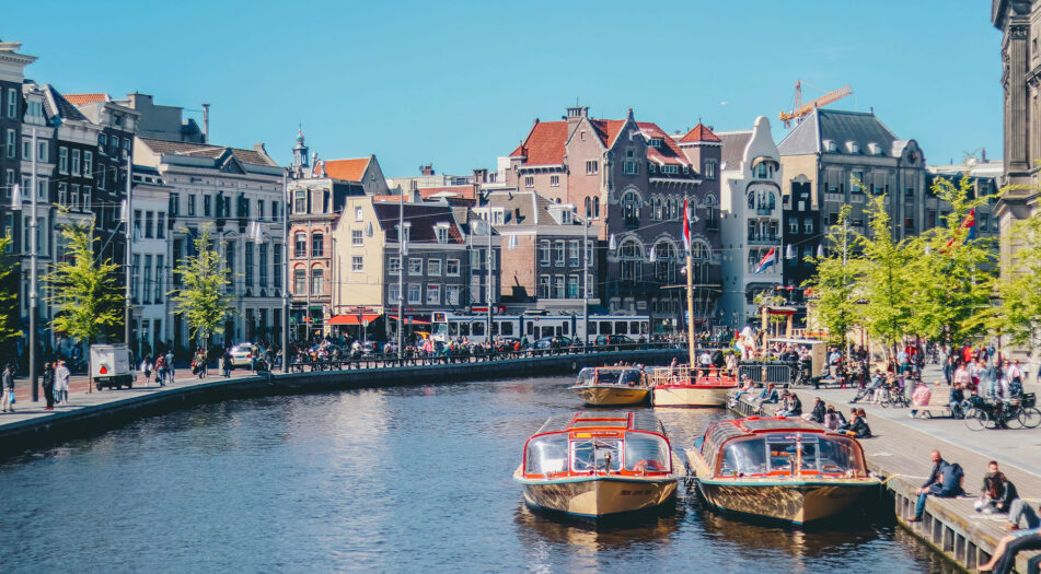 A scenic canal view in Amsterdam with two docked canal boats, traditional Dutch buildings with gabled roofs in the background, and people relaxing along the water’s edge.