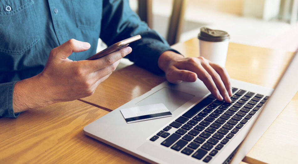 A person sits at a wooden table using a laptop, holding a smartphone in one hand and typing with the other.